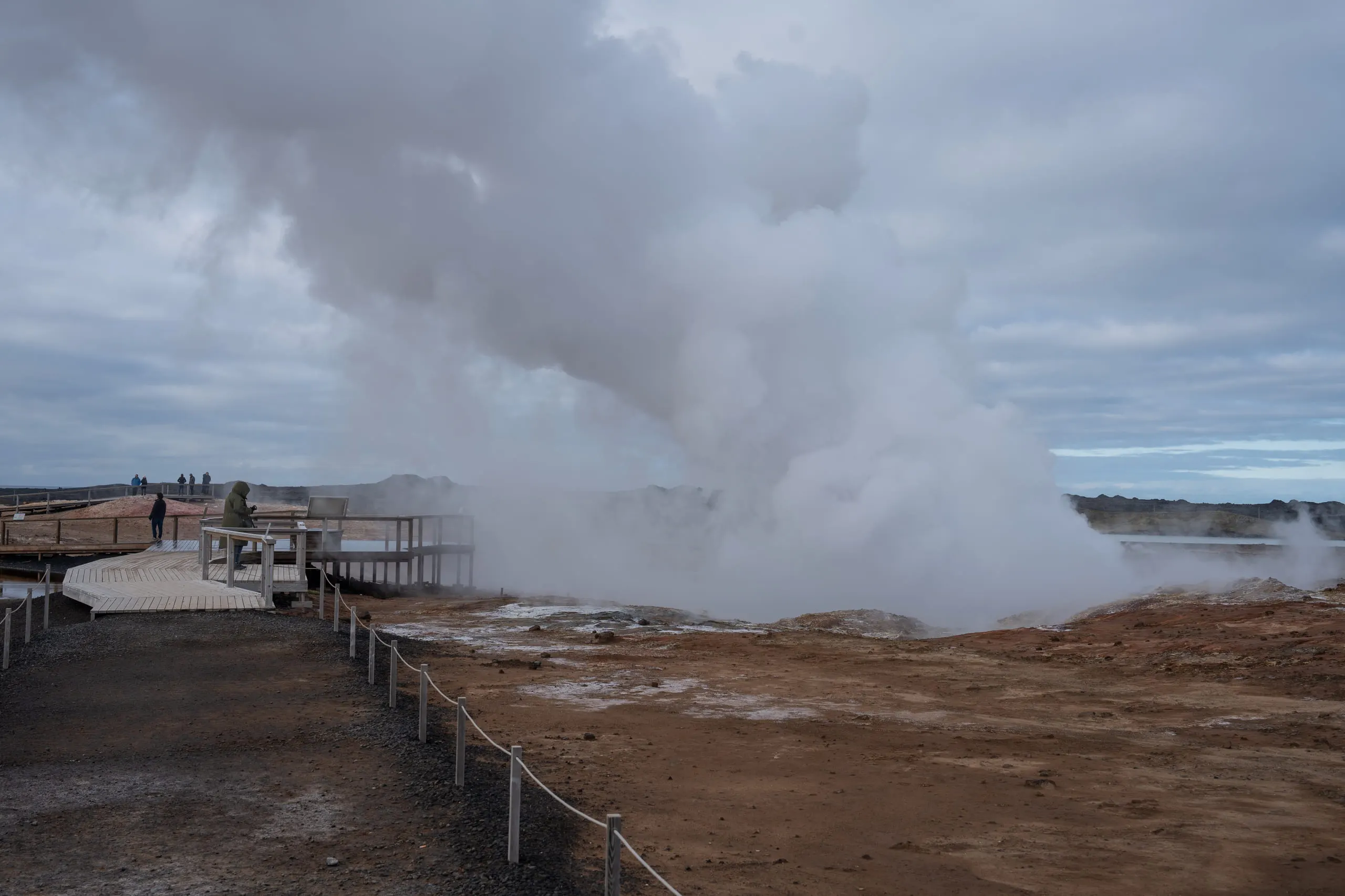 A wooden boardwalk crossing a steaming geothermal field under heavy grey cloud.