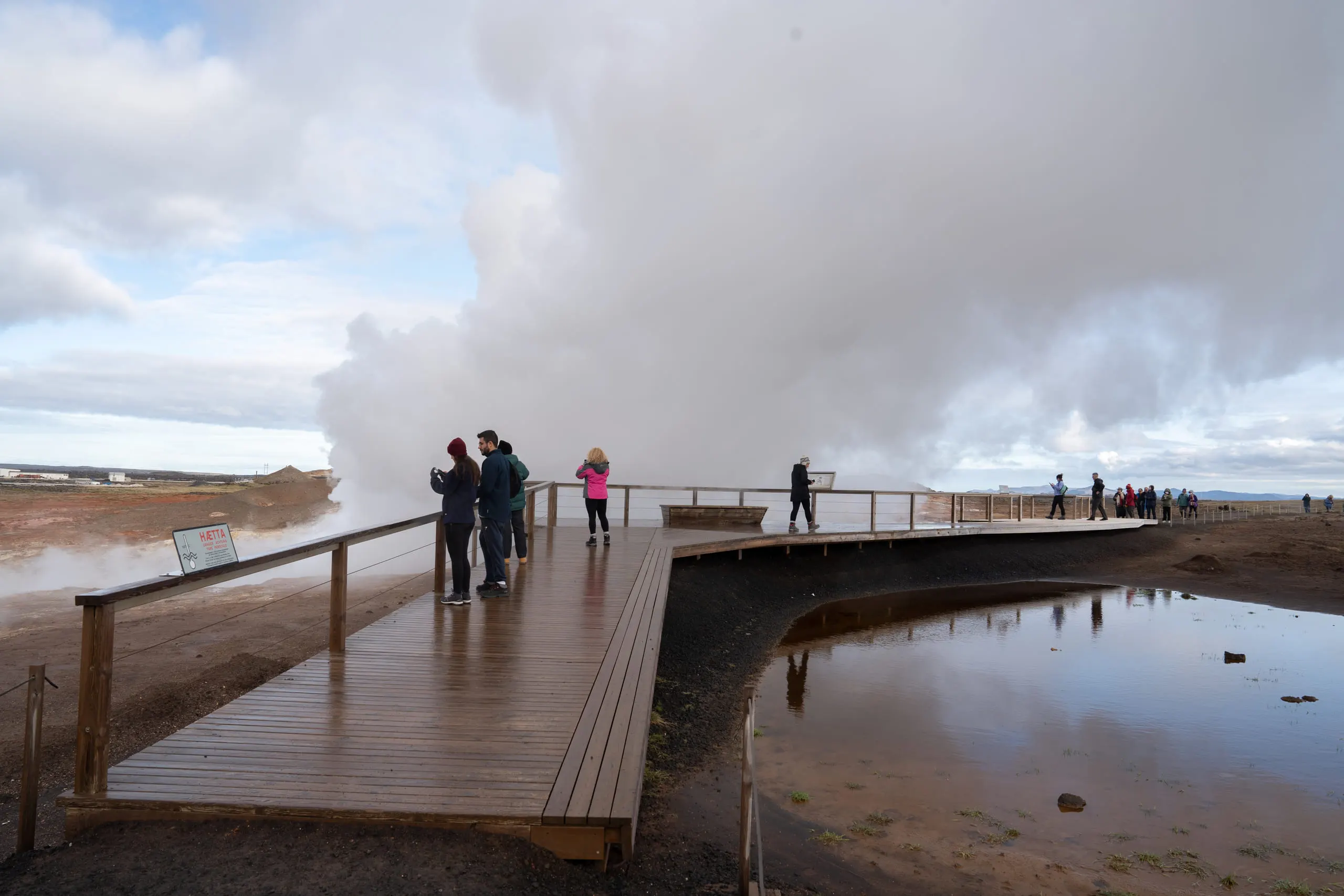 People walking a wet boardwalk with steam billowing behind them, reflected in standing water.