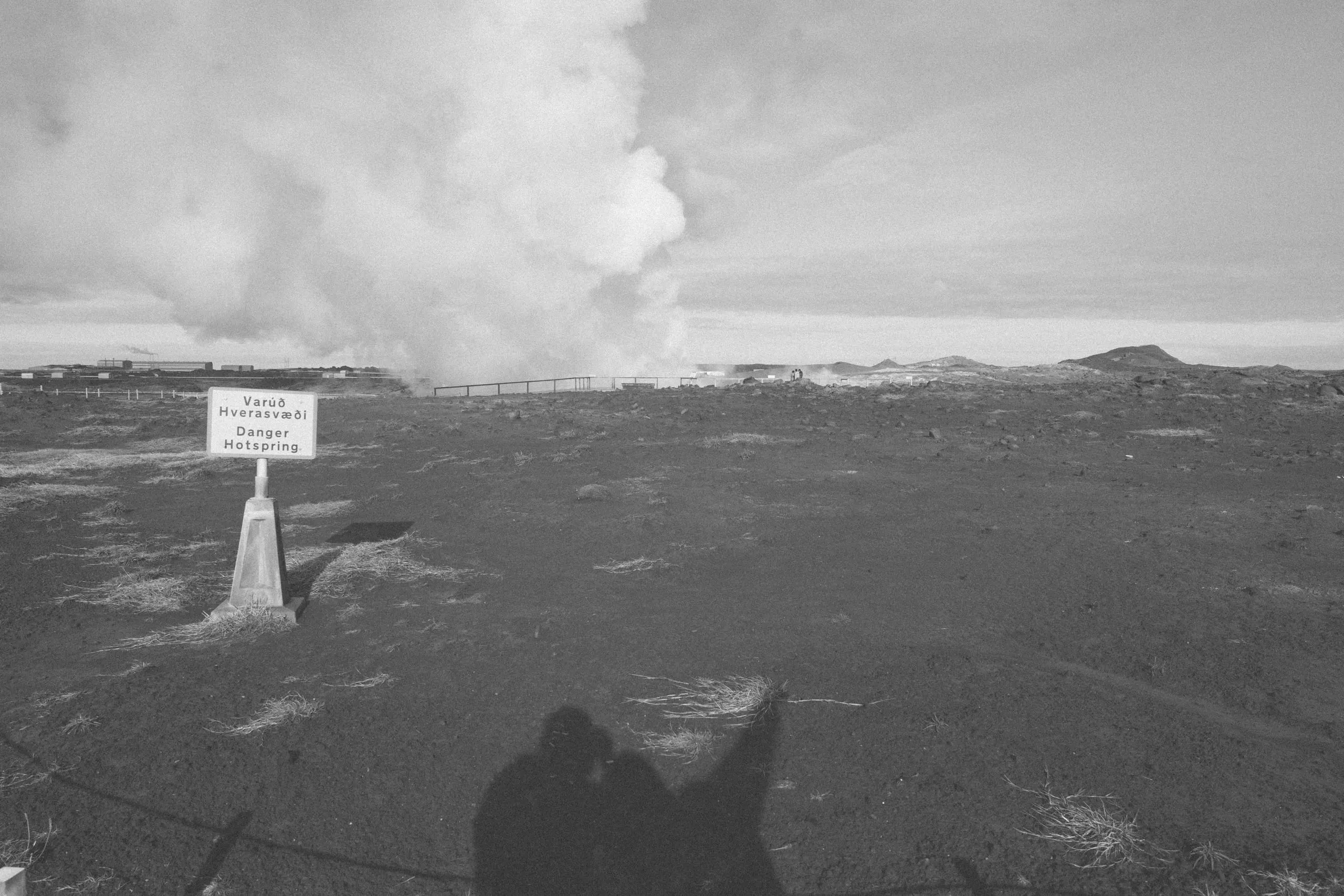 Black and white view across a barren steaming field, the photographer's shadow stretching into the foreground.