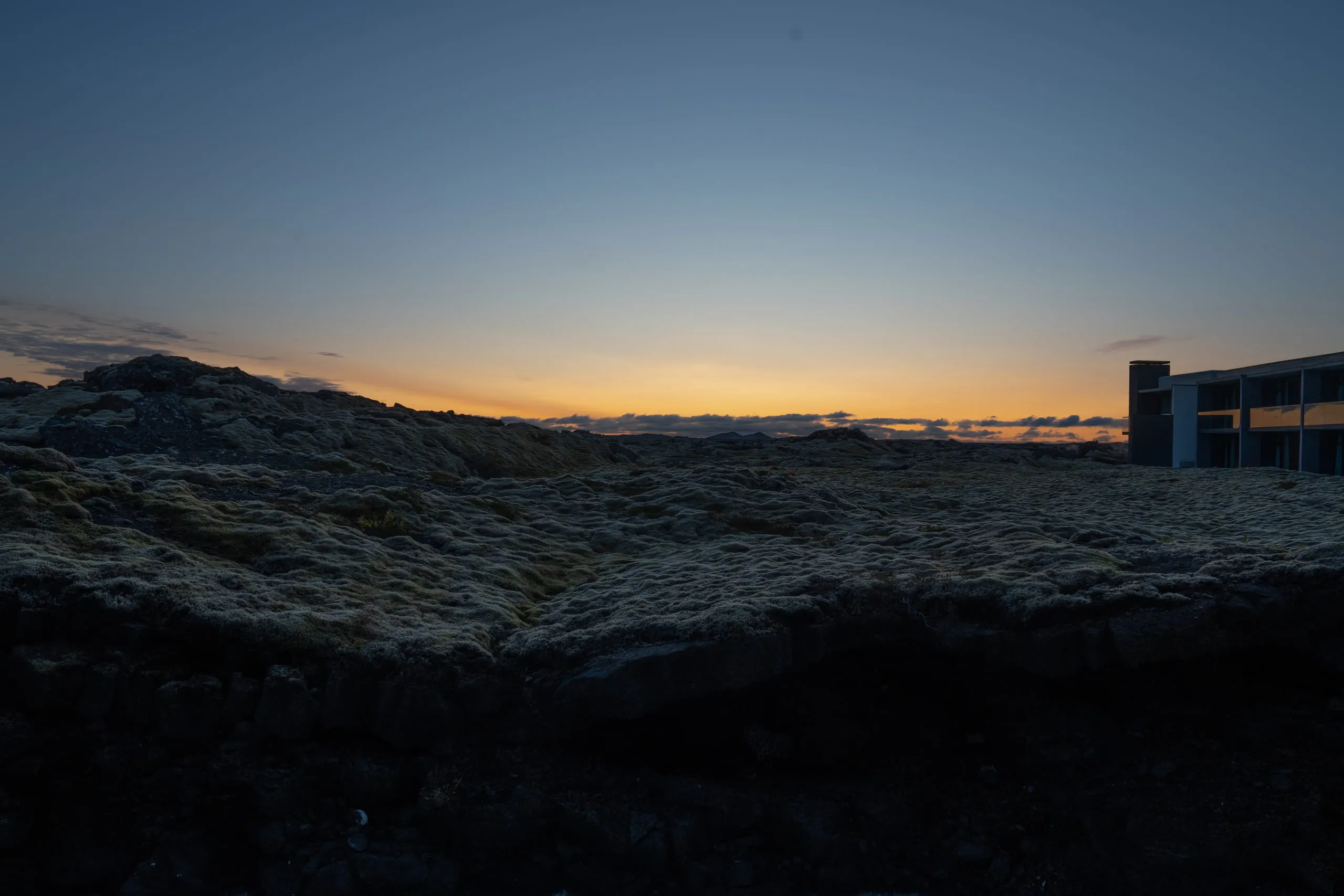 A dark lava field stretching to the horizon at dusk, a low lit building at the right edge.