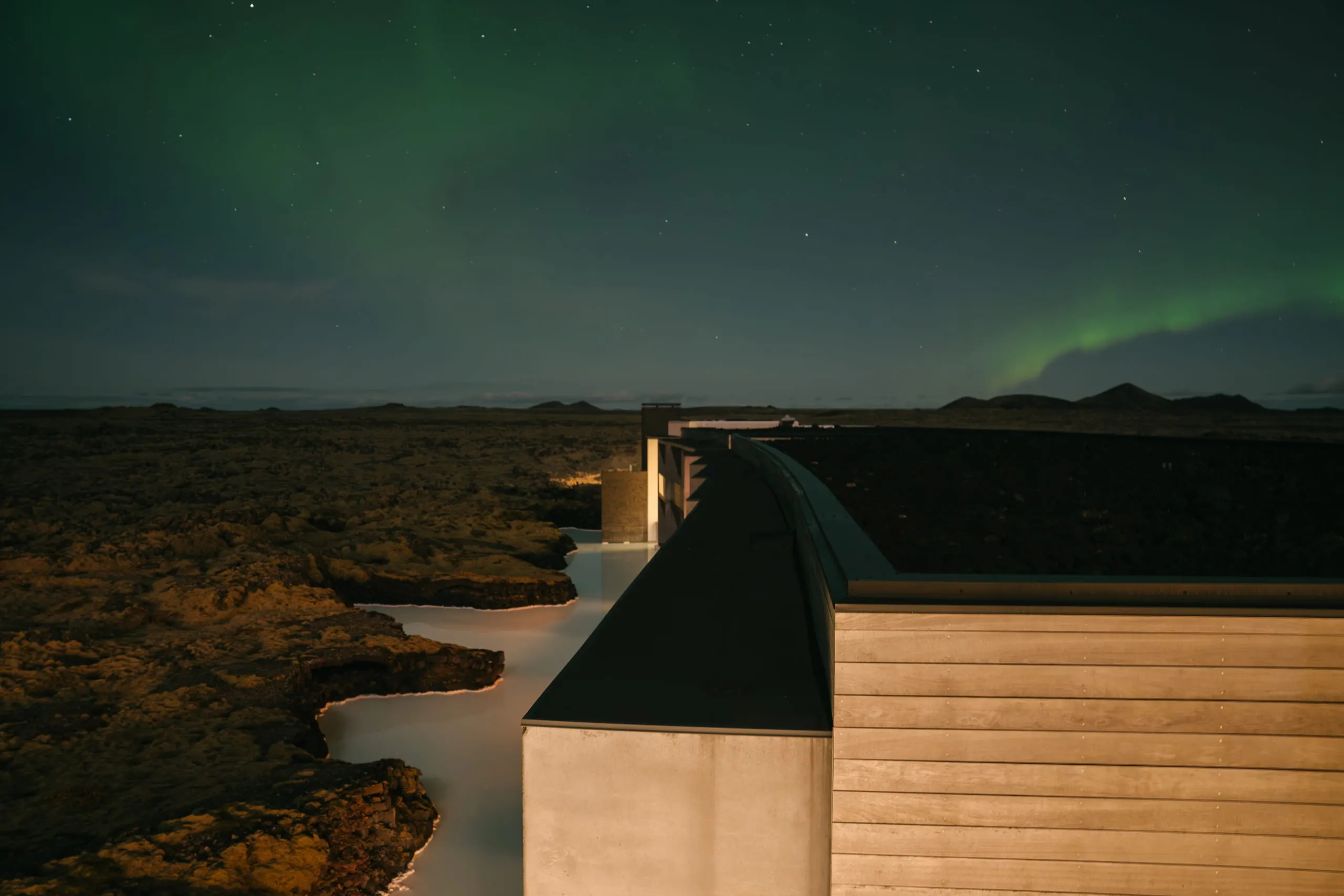 Green aurora arcing over a small cabin with warm lit windows, beside a dark stream.