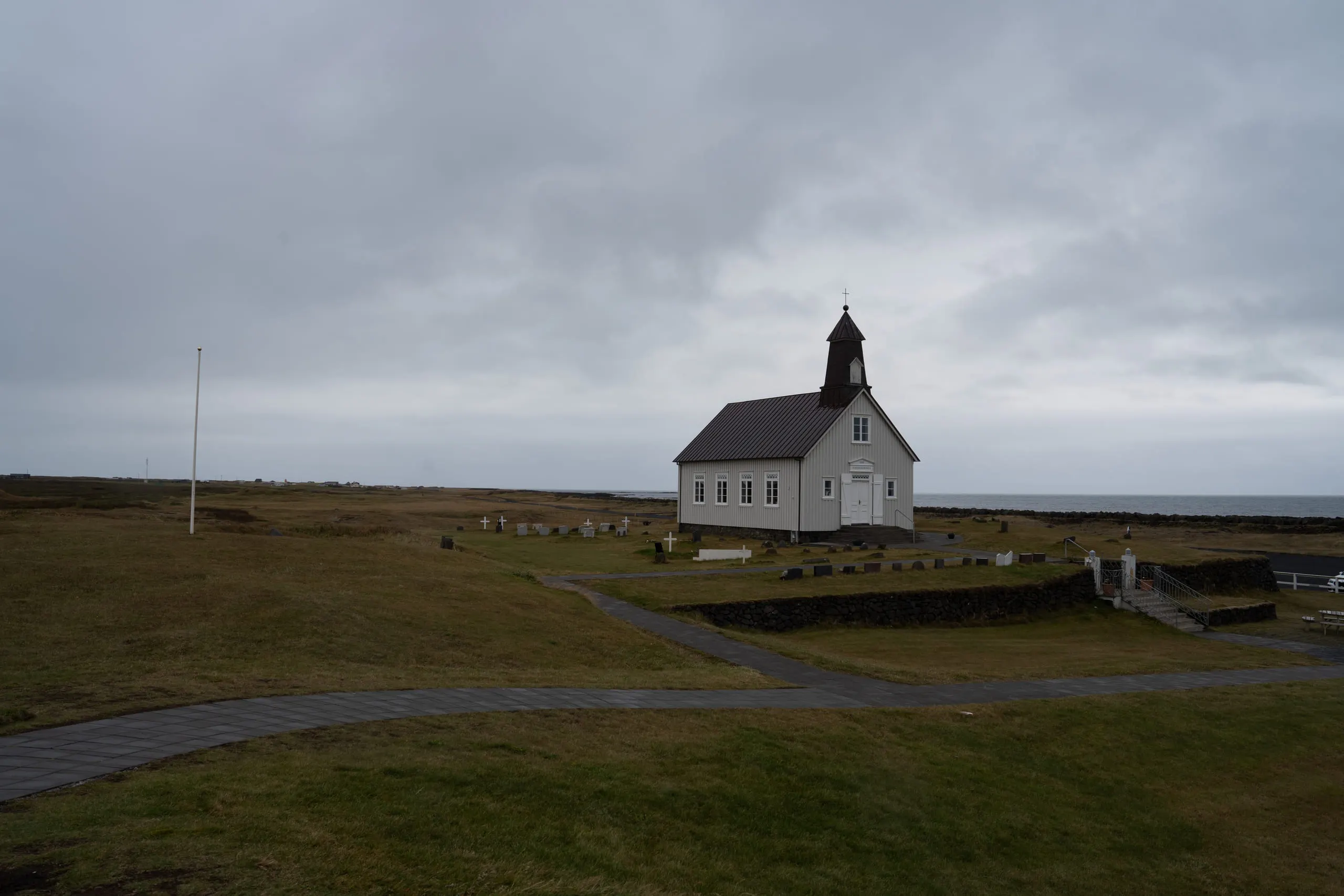 A small white church with a black roof standing alone on a wide green plain under grey sky.