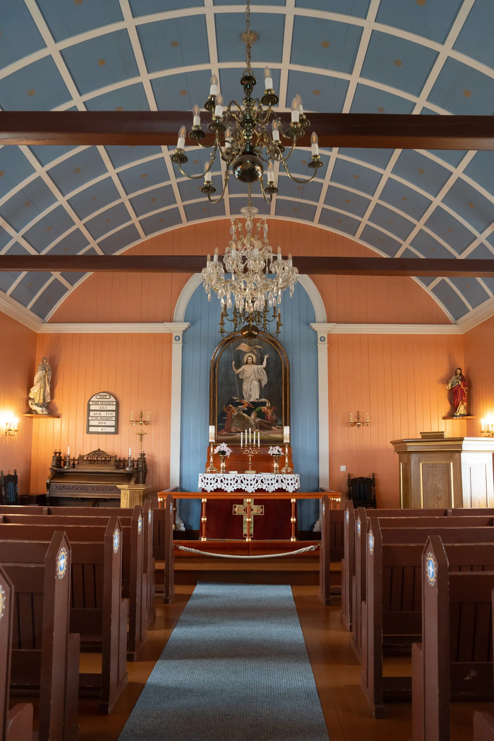 The interior of a small church looking down the aisle to a blue and gold altar beneath a chandelier.