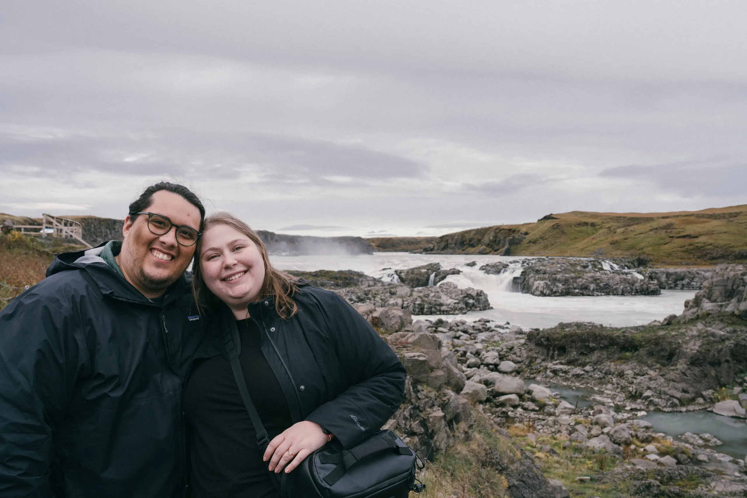 Adrian and Kylie standing together on a gravel path, a wide braided river running behind them.