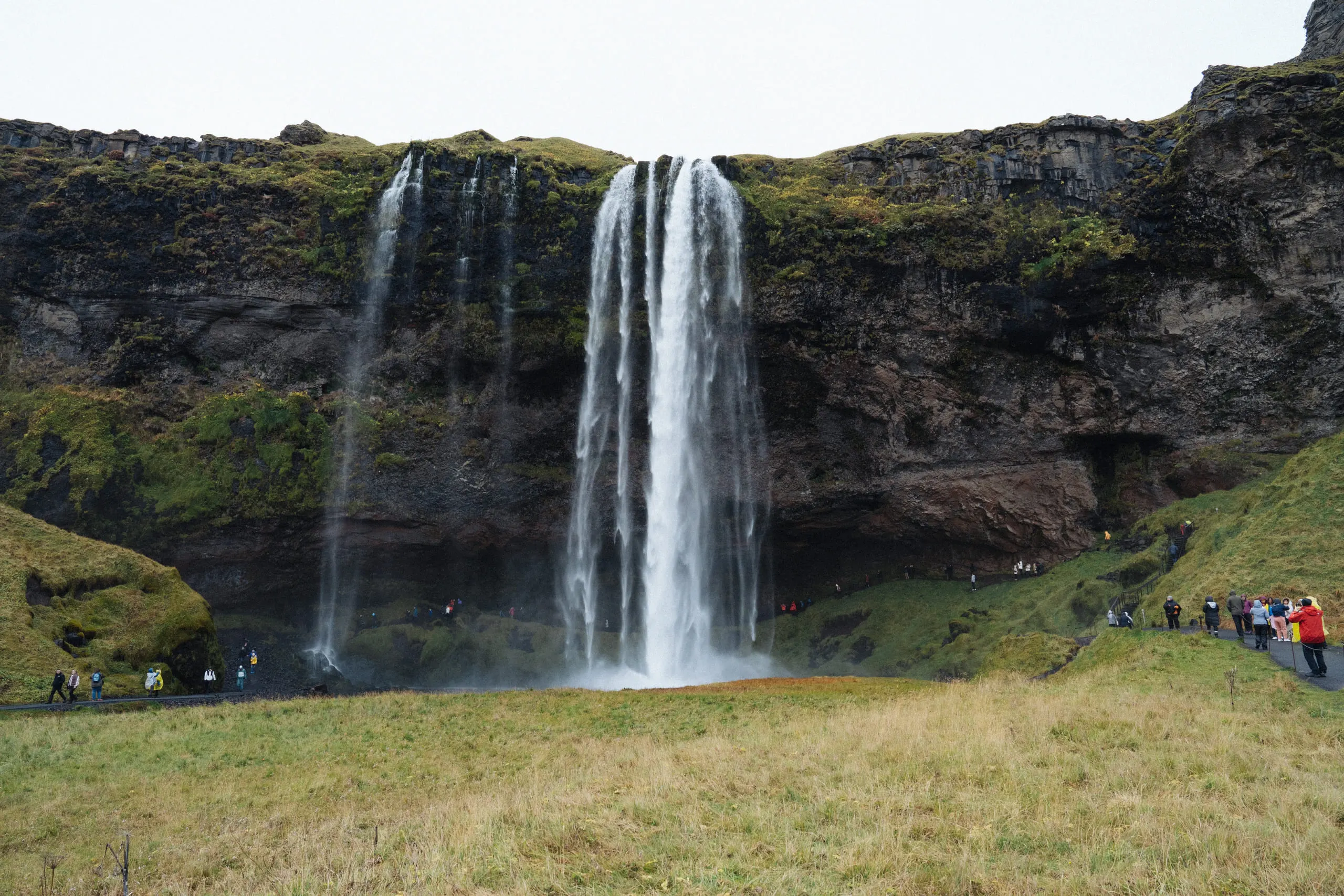 Seljalandsfoss falling in a single ribbon over a green cliff, with people small at its base.