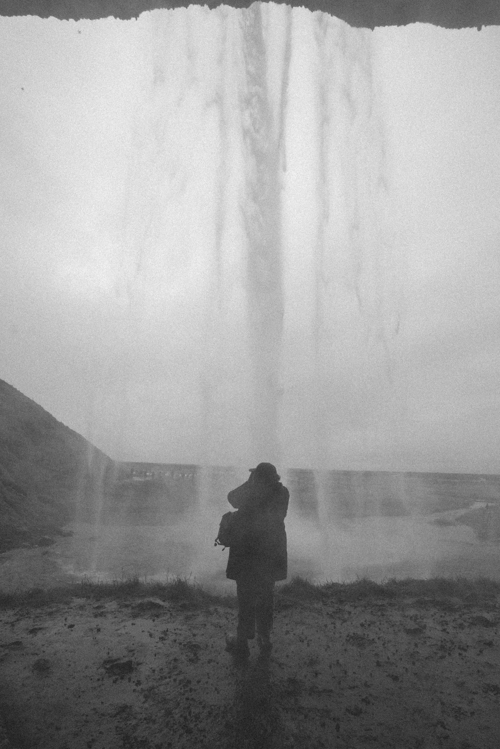 Black and white view from behind a waterfall, a lone figure silhouetted against the sheet of falling water.