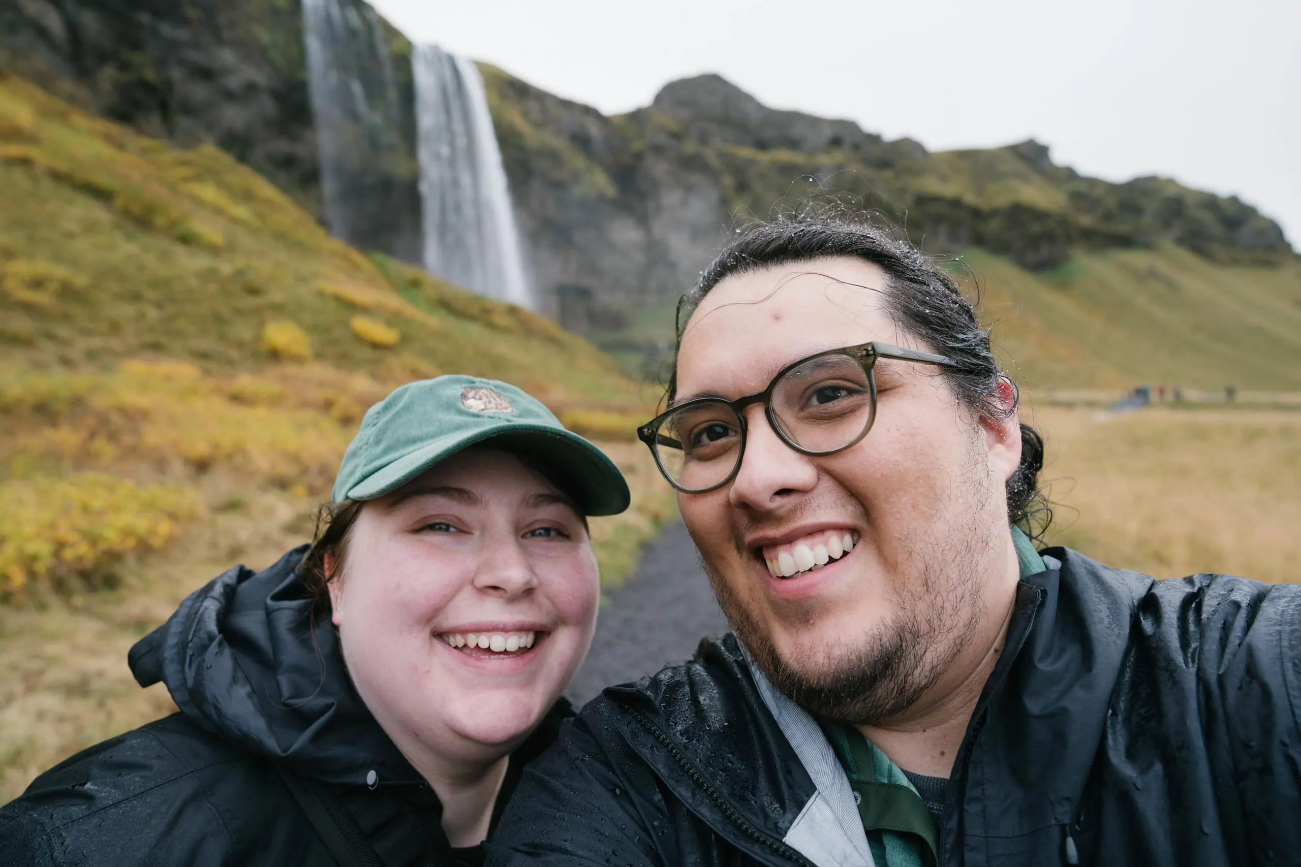 Adrian and Kylie grinning in a close selfie, a green hillside and waterfall behind them.