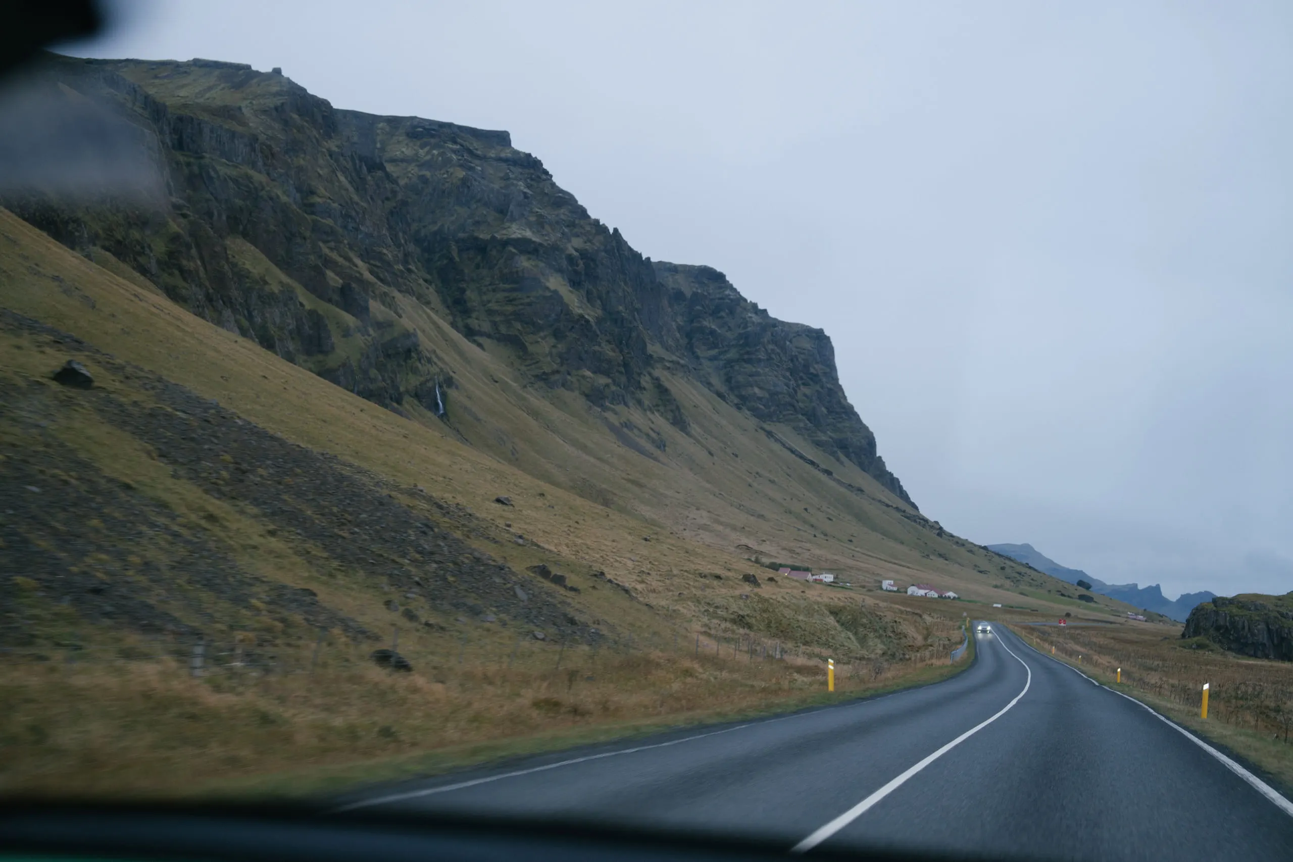 A road curving away beneath a long green and grey escarpment, seen through a car windscreen.