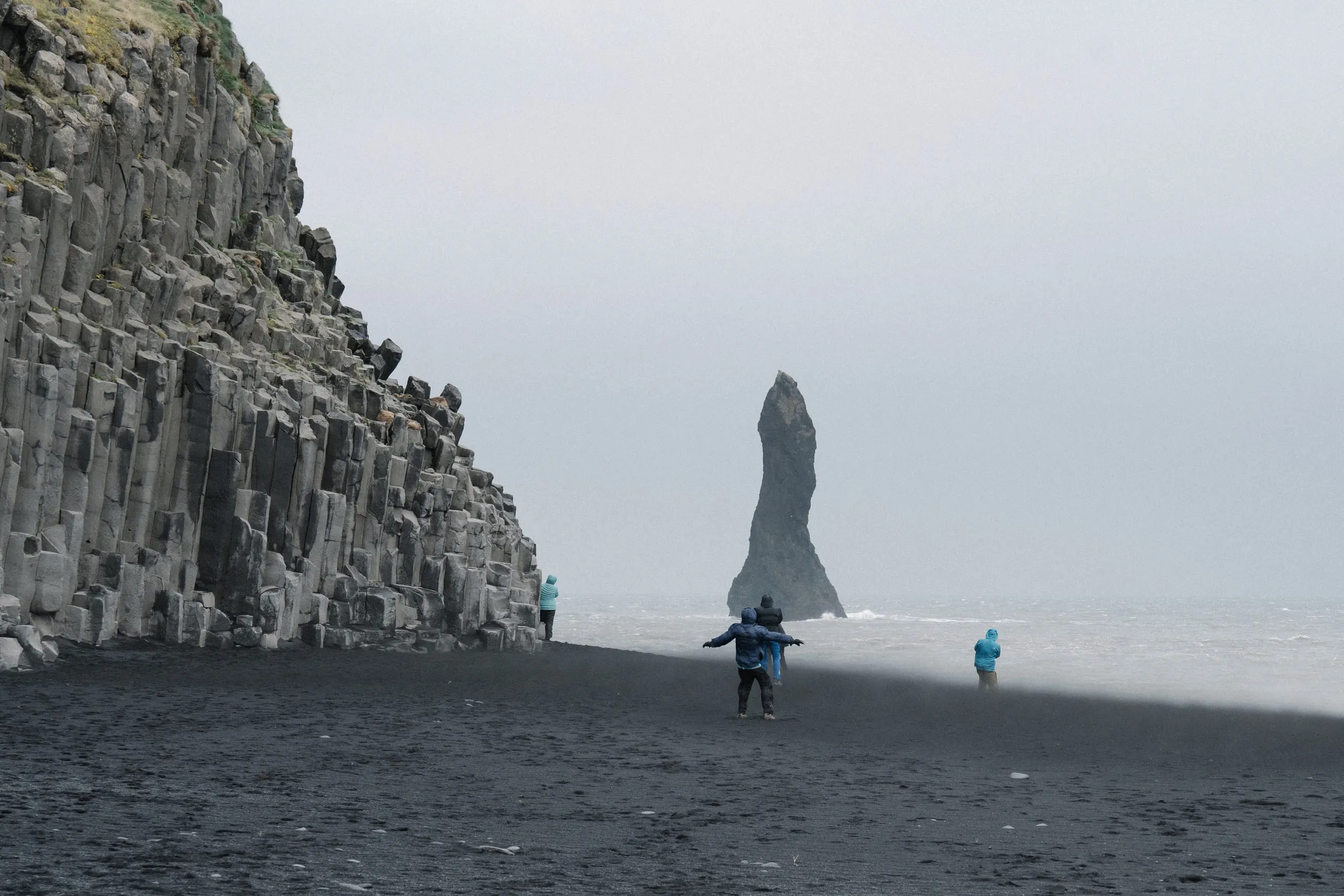 Stacked basalt columns along a black sand beach, a jagged sea stack rising offshore.