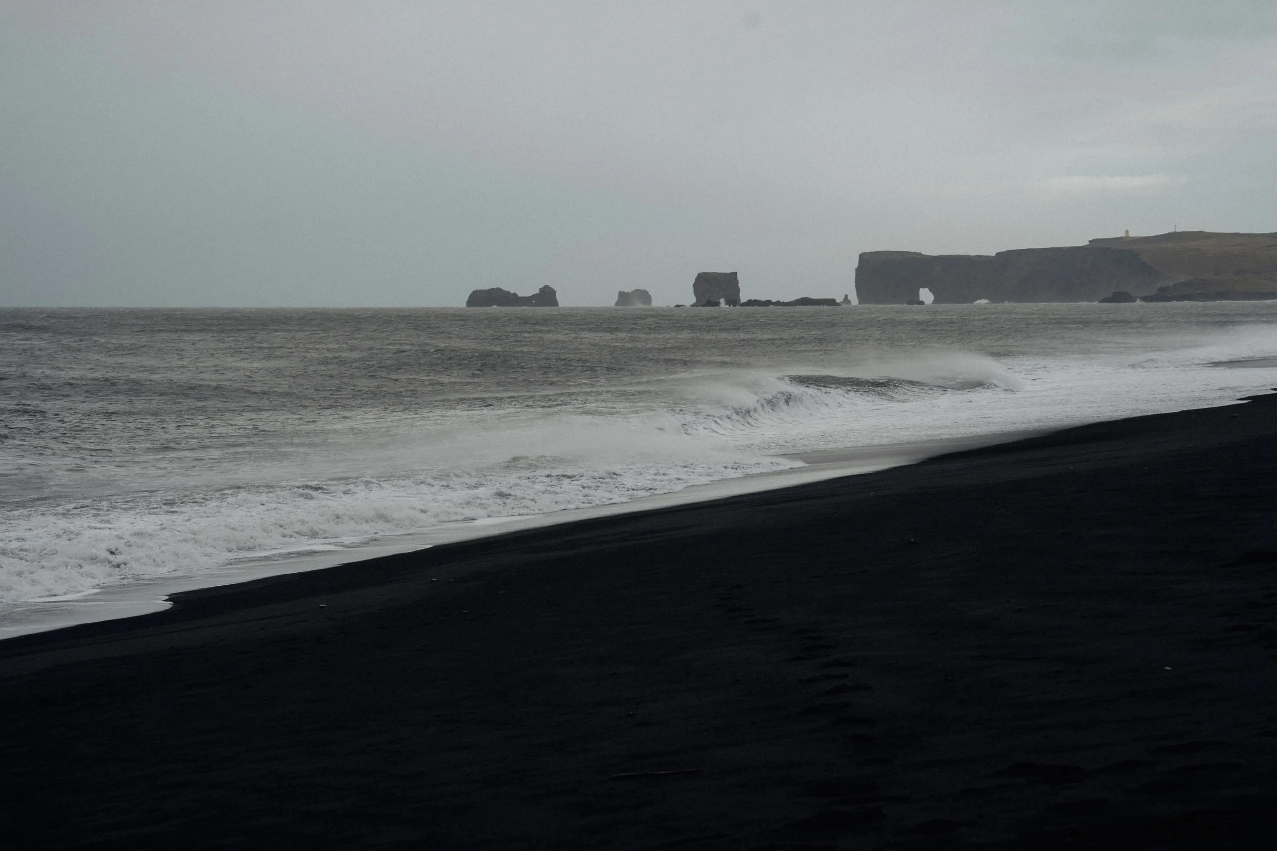 A diagonal sweep of black sand meeting pale surf, a dark headland low on the horizon.