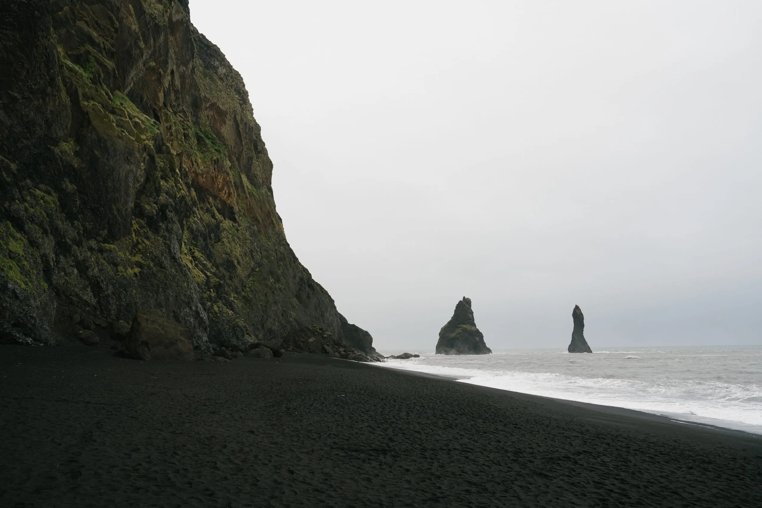 Sea stacks standing offshore beyond a steep cliff face and a black sand beach.