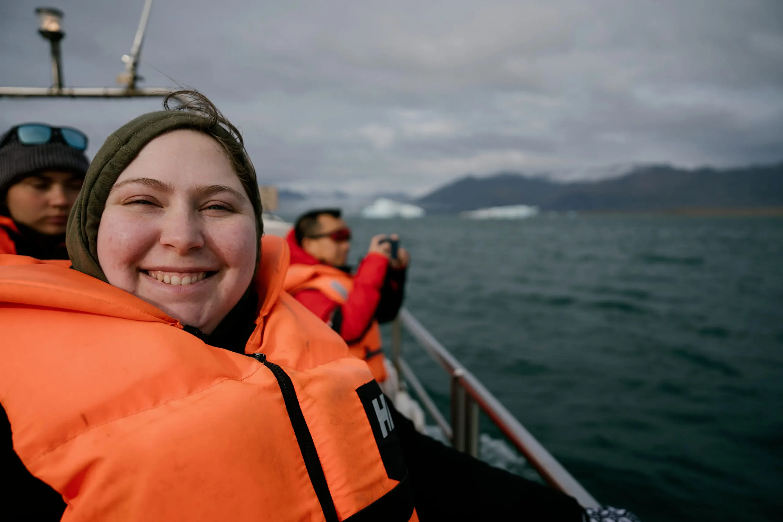 Kylie beaming in an orange life vest aboard a small boat, glacial water and low hills behind her.