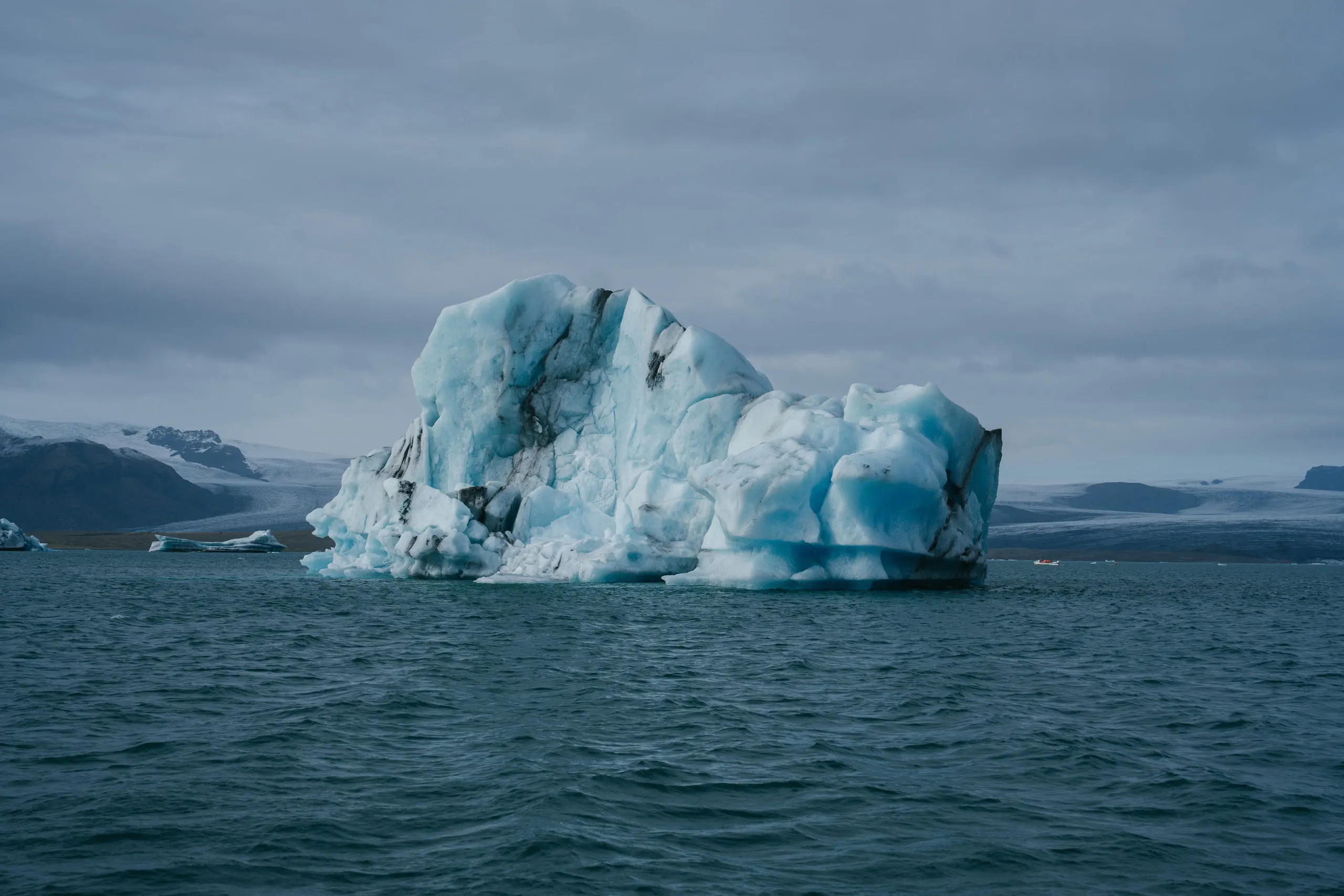 A blue and white iceberg floating alone in a glacial lagoon, low mountains behind it.