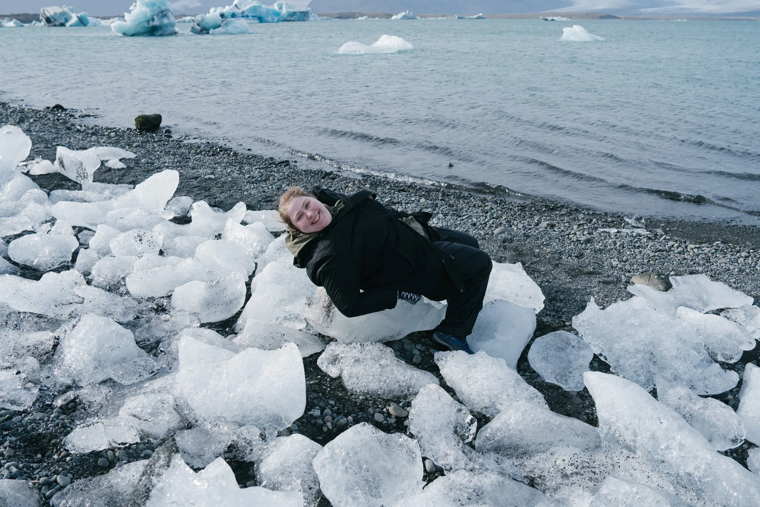 Kylie lying back among chunks of clear glacial ice scattered across a black sand beach.