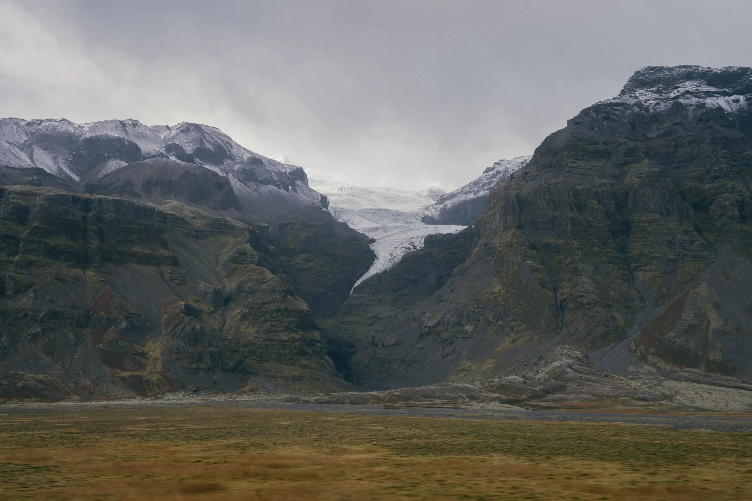 A glacier tongue spilling between dark mountains down toward a grey plain.