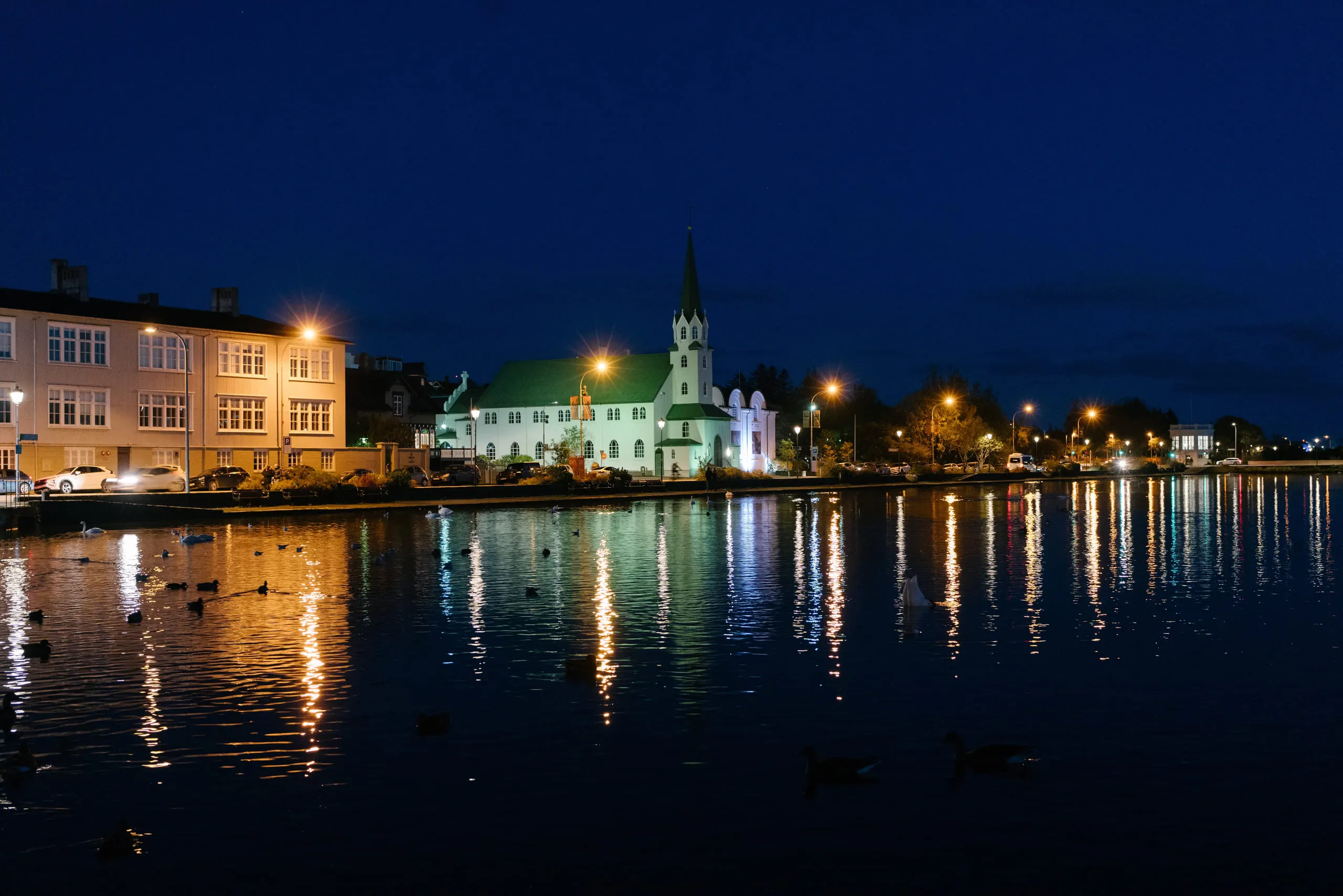 Waterfront buildings lit at blue hour, their reflections running down the still surface of a pond.