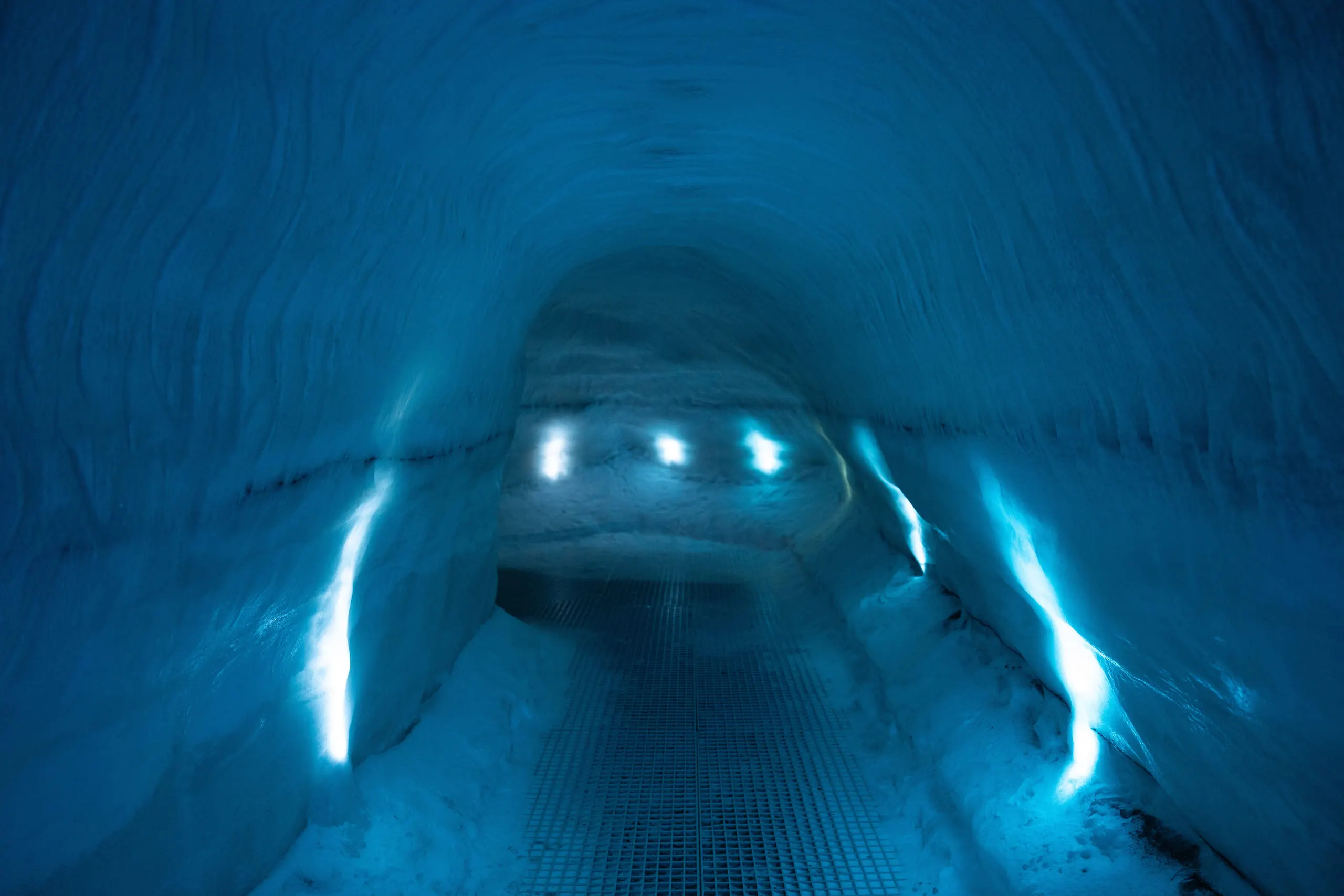 A tunnel carved through ice and lit deep blue, curving away out of sight.
