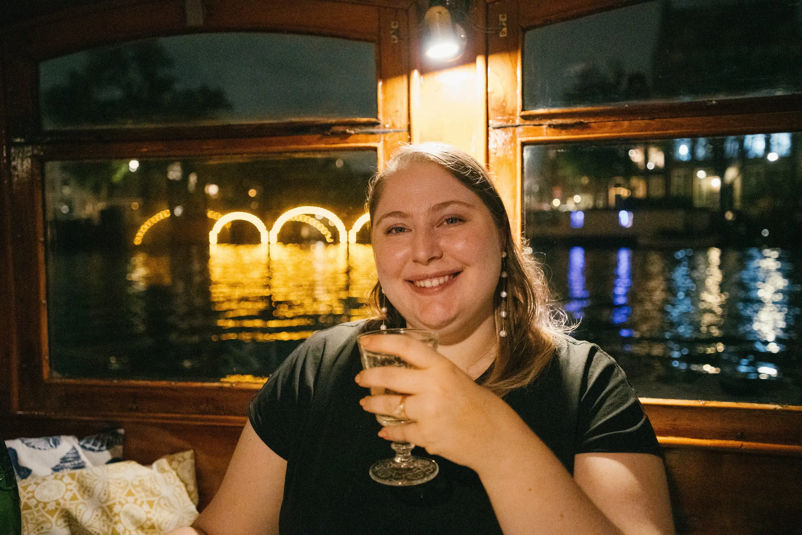 Kylie raising a glass at a table on the boat, canal lights strung in the window behind her.