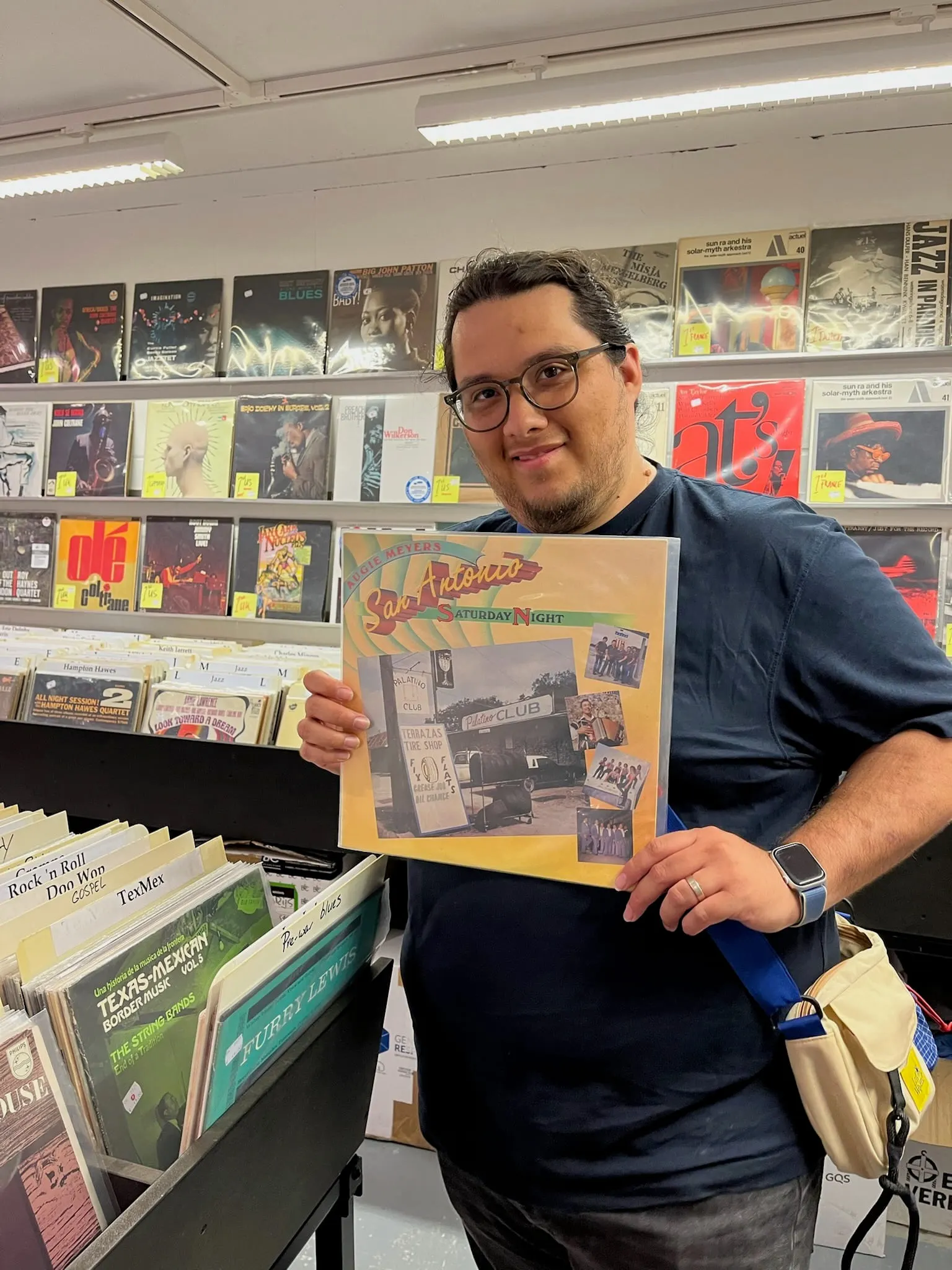 Adrian holding up a record in a shop, racks of sleeves filling the wall behind him.