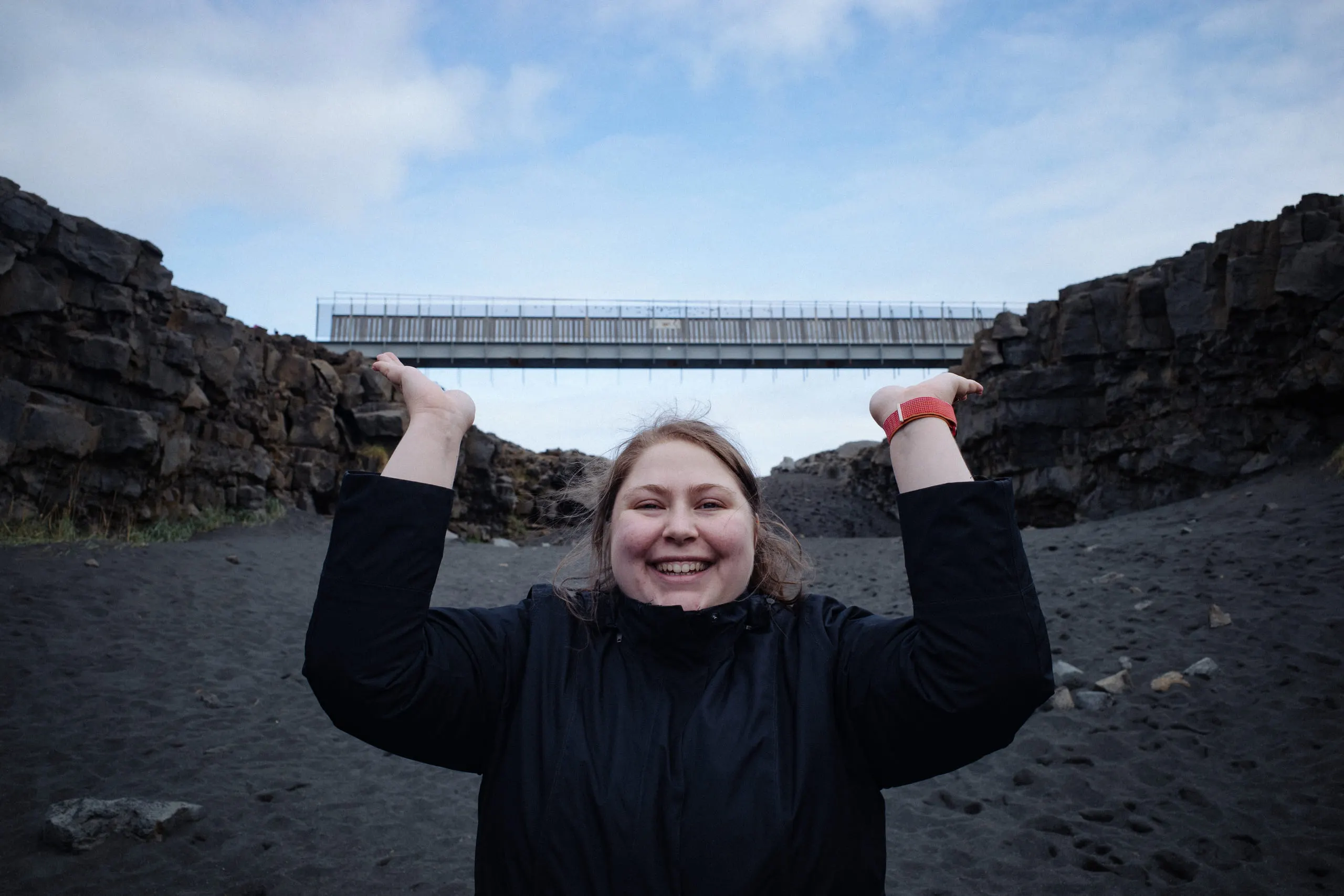 Kylie standing with both arms raised beneath a footbridge spanning a rift in black volcanic gravel.