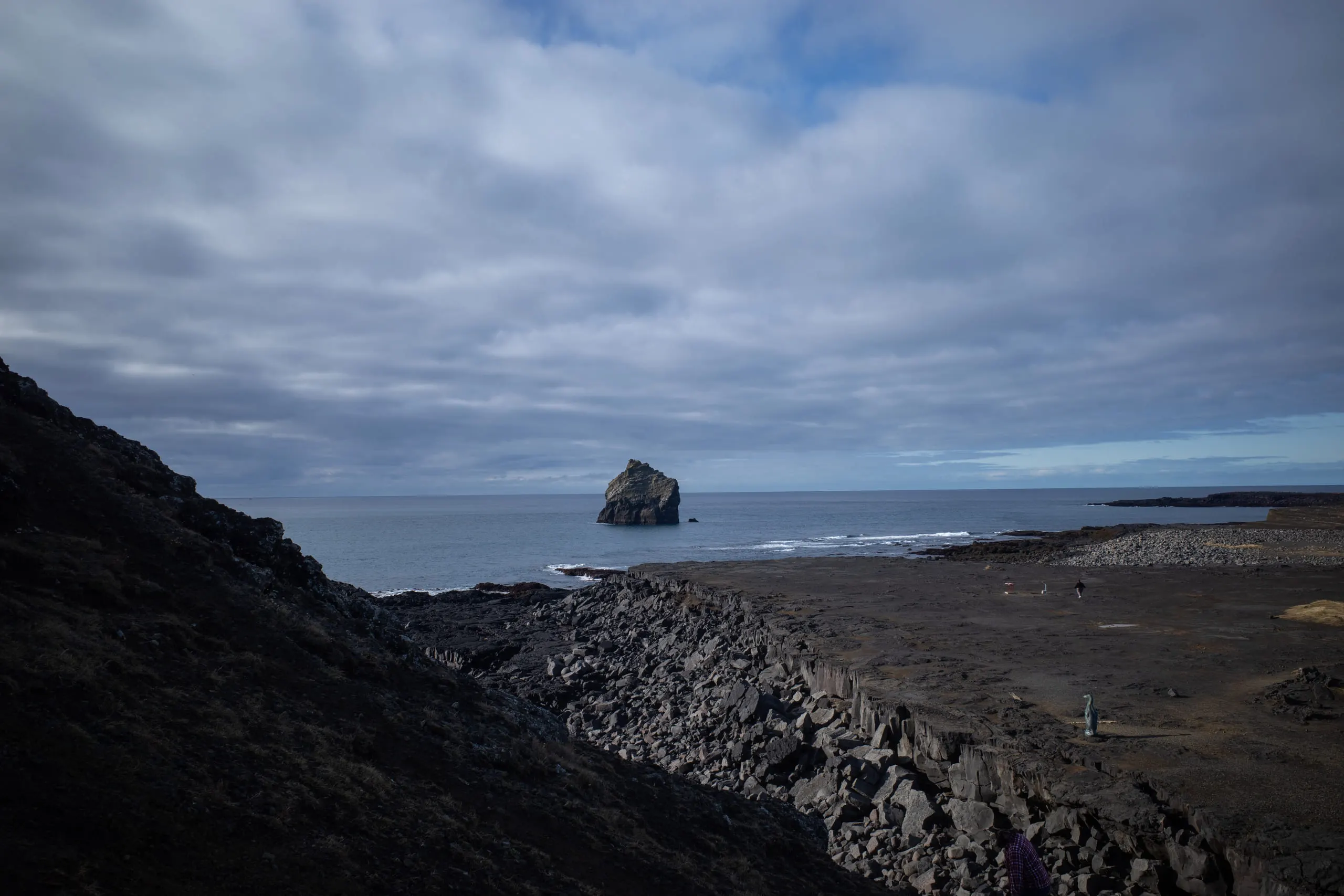 A lone sea stack offshore, seen across a boulder-strewn black shoreline under low grey cloud.