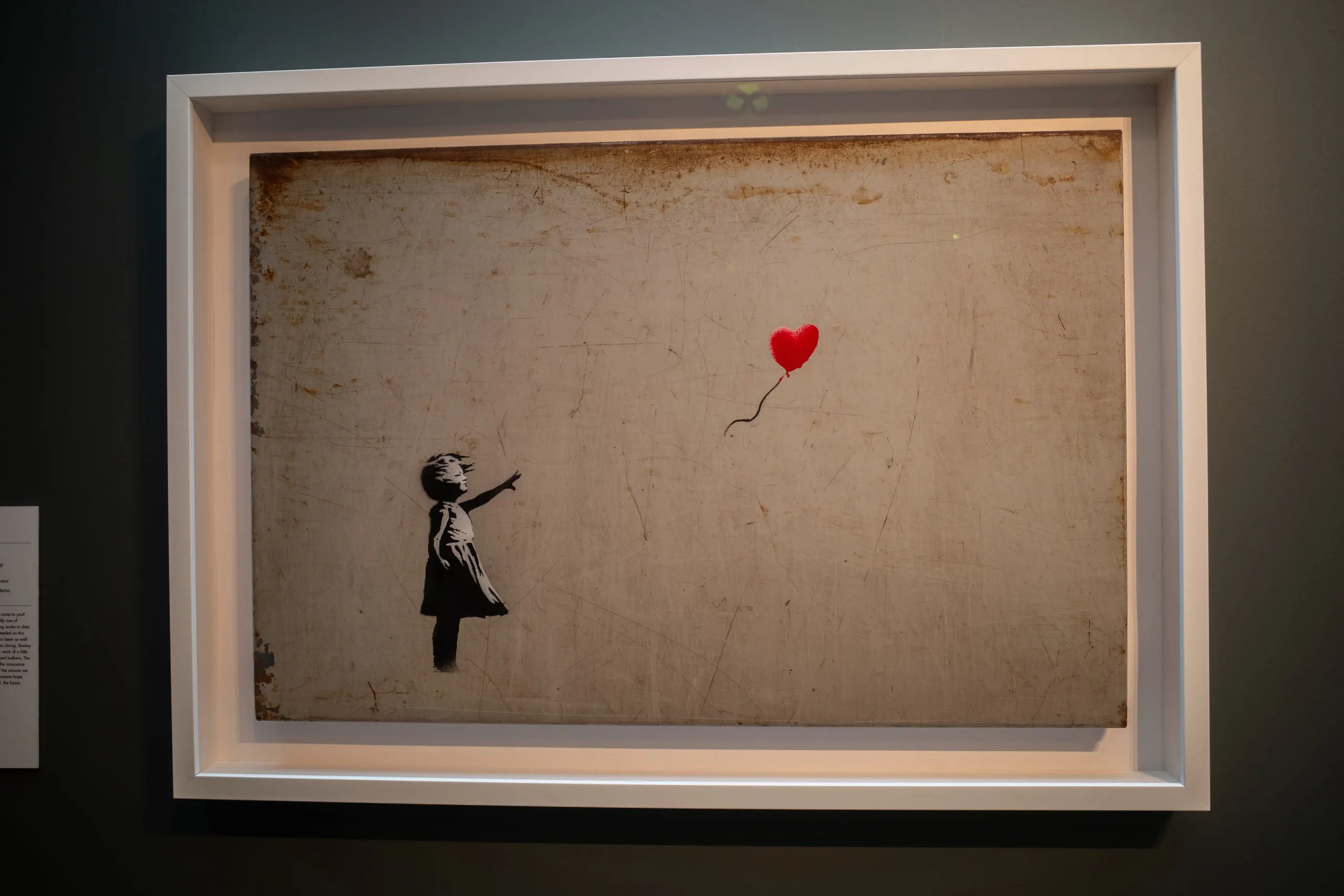 Banksy's Girl with Balloon framed on a dark gallery wall, the red balloon the only colour in the frame.